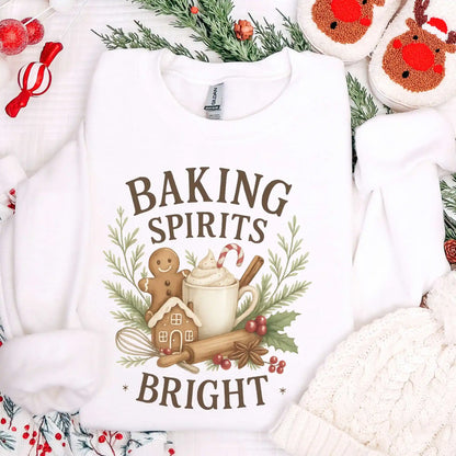 White-colored Beacon & Bramble Christmas sweatshirt featuring the phrase ‘Baking Spirits Bright’ above a festive design with a gingerbread man, mug of cocoa topped with whipped cream and a candy cane, cinnamon sticks, greenery, and holly berries. Surrounded by cozy holiday decor including a knit hat, reindeer slippers, and greenery accents.
