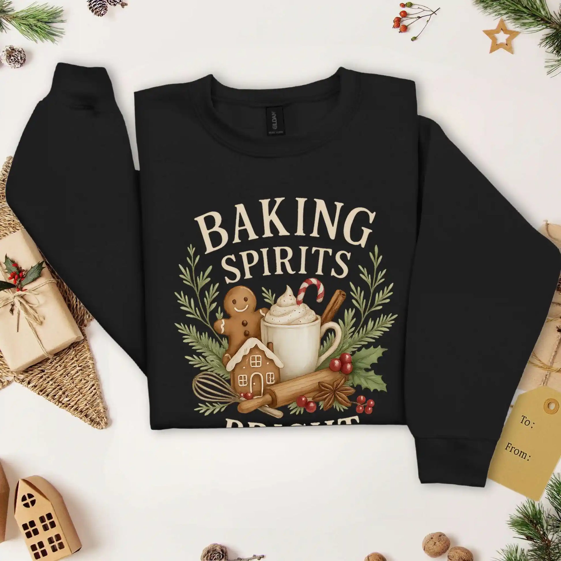 Black colored Beacon & Bramble Christmas sweatshirt featuring the phrase ‘Baking Spirits Bright’ above a festive design with a gingerbread man, mug of cocoa topped with whipped cream and a candy cane, cinnamon sticks, greenery, and holly berries. Surrounded by cozy holiday decor and greenery accents.