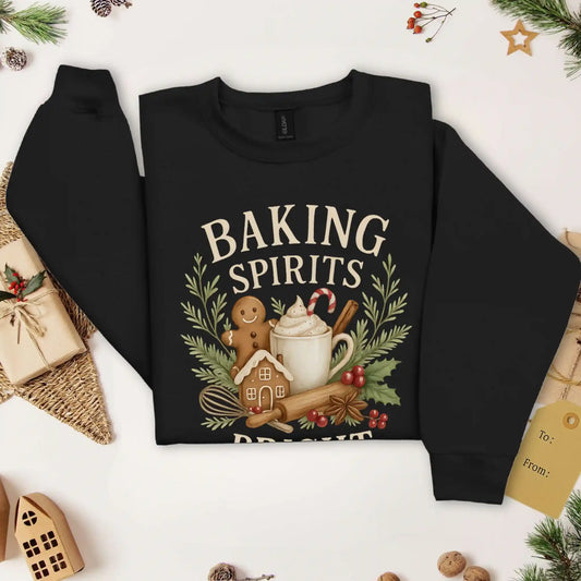 Black colored Beacon & Bramble Christmas sweatshirt featuring the phrase ‘Baking Spirits Bright’ above a festive design with a gingerbread man, mug of cocoa topped with whipped cream and a candy cane, cinnamon sticks, greenery, and holly berries. Surrounded by cozy holiday decor and greenery accents.