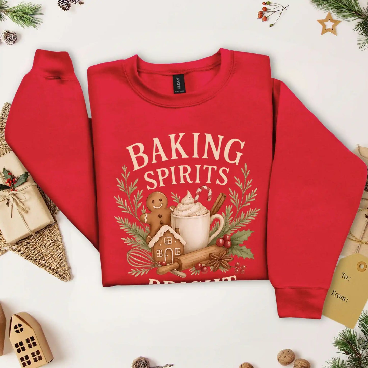 Cardinal red-colored Beacon & Bramble Christmas sweatshirt featuring the phrase ‘Baking Spirits Bright’ above a festive design with a gingerbread man, mug of cocoa topped with whipped cream and a candy cane, cinnamon sticks, greenery, and holly berries. Surrounded by cozy holiday decor and greenery accents.