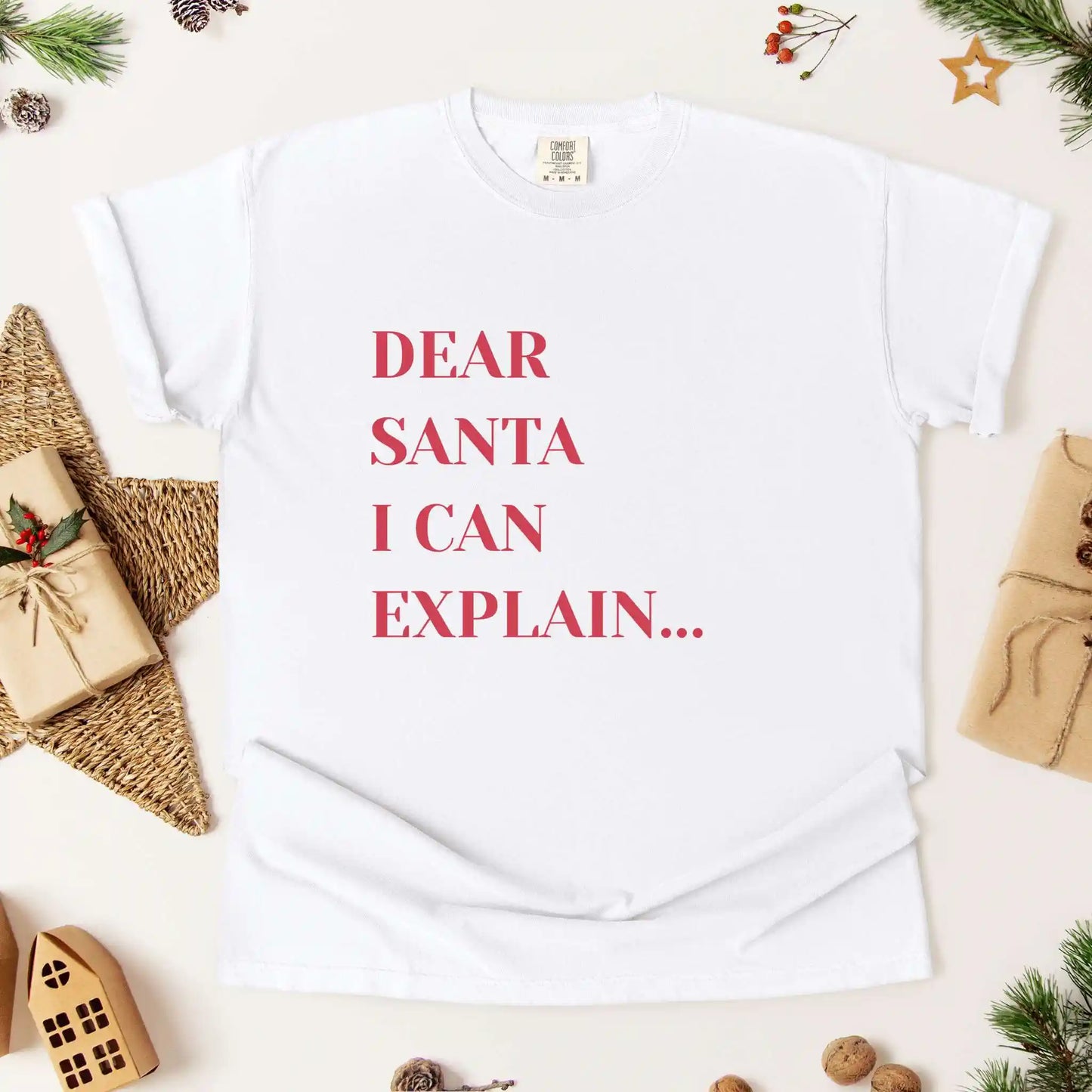 White t-shirt with 'Dear Santa I can explain' text on a festive background with Christmas decorations.