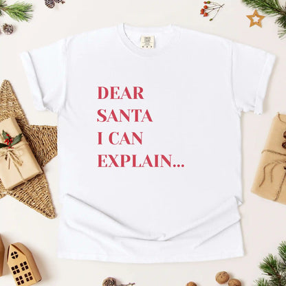 White t-shirt with 'Dear Santa I can explain' text on a festive background with Christmas decorations.