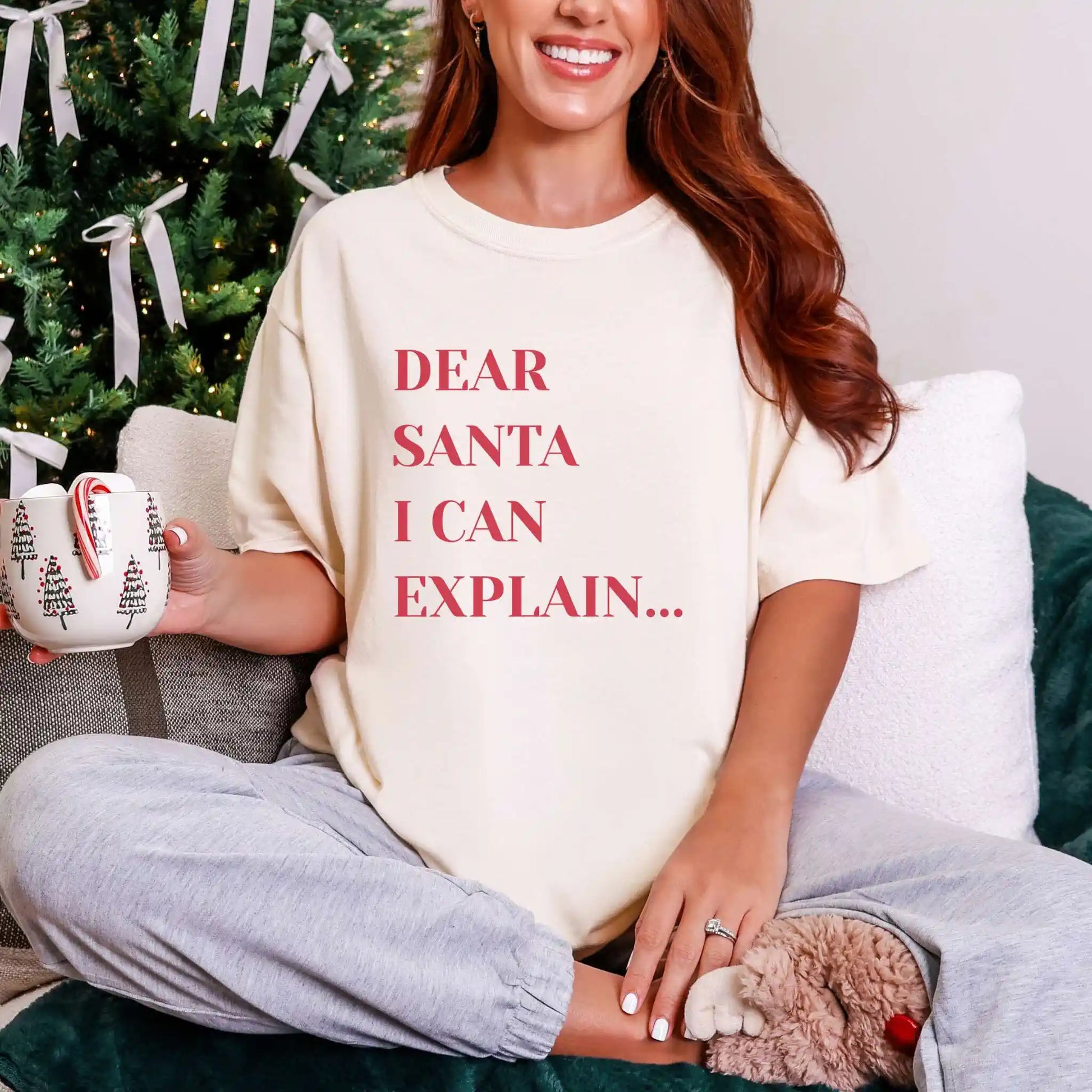 Person wearing a shirt with 'Dear Santa I can explain' text, holding a mug with candy cane, sitting in front of a Christmas tree.