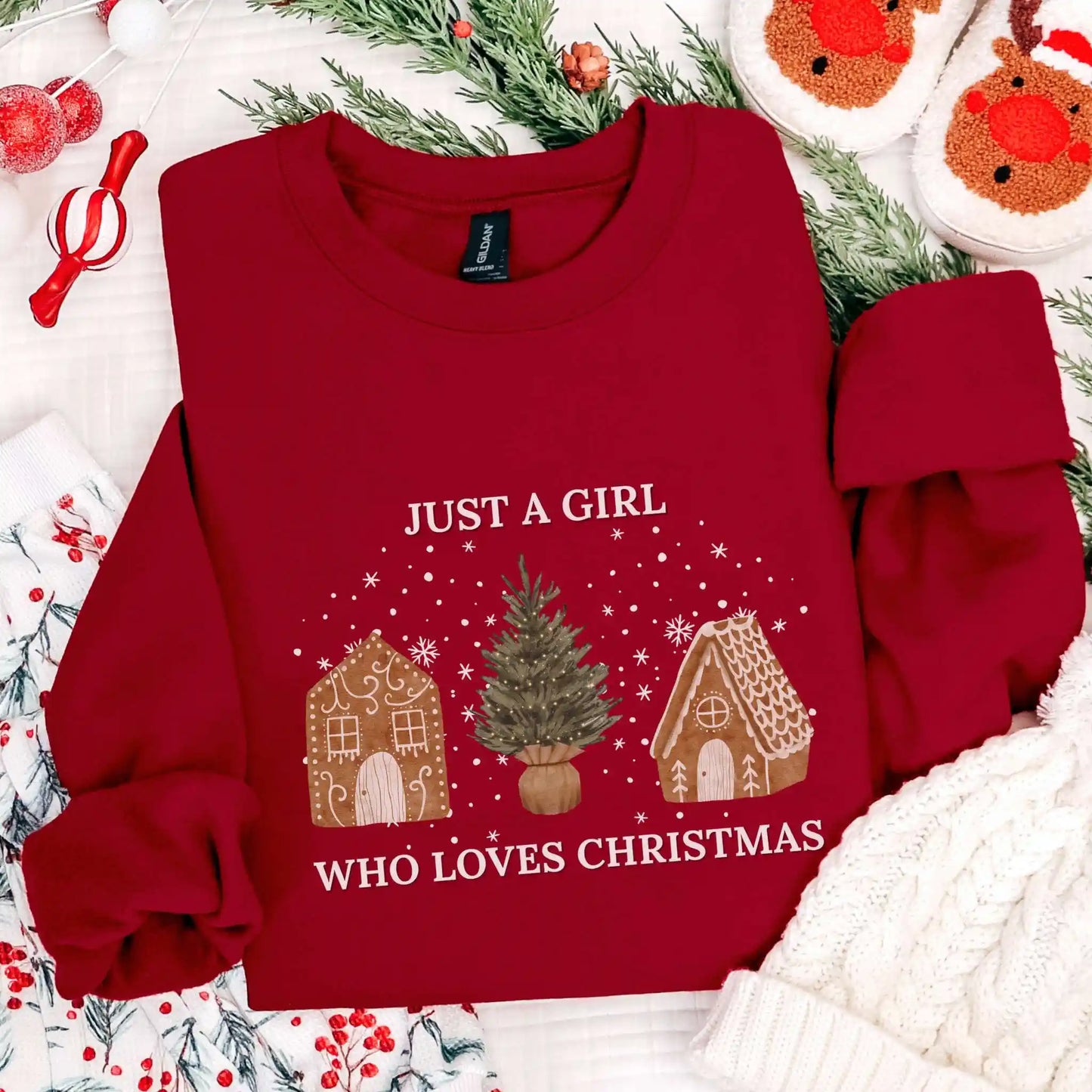 Garnet red-colored Beacon & Bramble Christmas sweatshirt featuring the phrase ‘Just a Girl’ above and ‘Who Loves Christmas’ below a festive design of two gingerbread houses and a Christmas tree, surrounded by cozy holiday decor including a knit hat, reindeer slippers, and greenery.