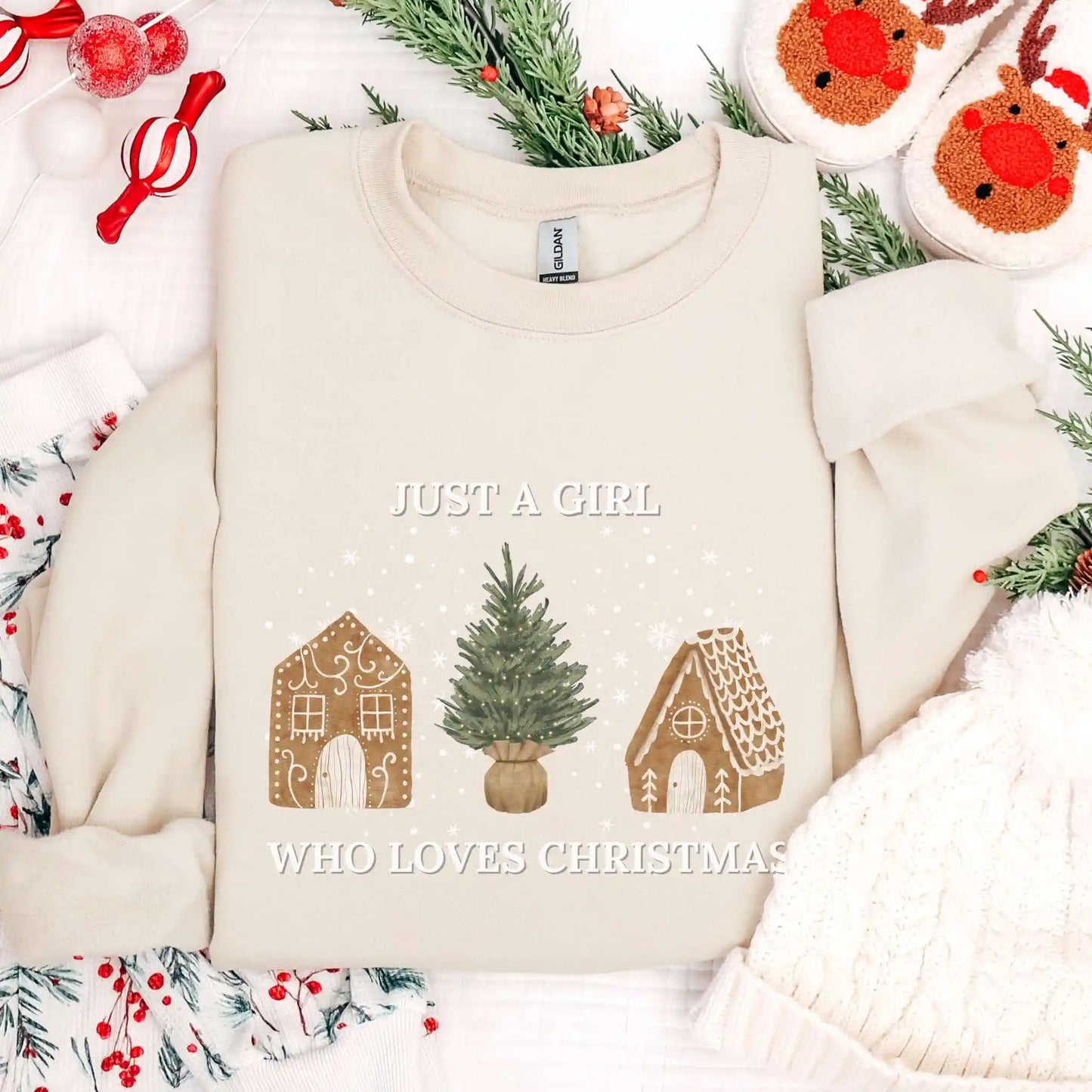 Creamy sand-colored Beacon & Bramble Christmas sweatshirt featuring the phrase ‘Just a Girl’ above and ‘Who Loves Christmas’ below a festive design of two gingerbread houses and a Christmas tree, surrounded by cozy holiday decor including a knit hat, reindeer slippers, and greenery.