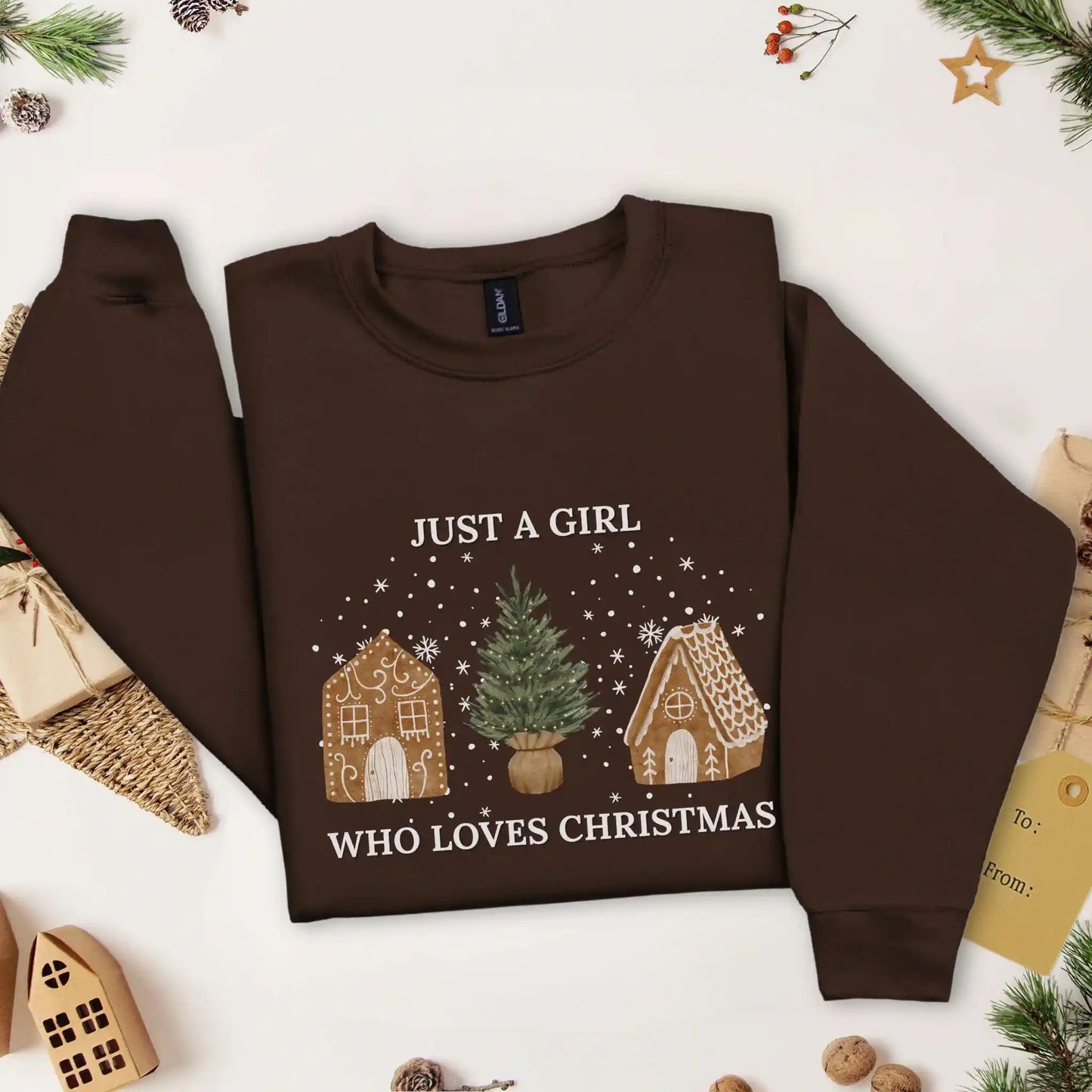 Dark Chocolate brown-colored Beacon & Bramble Christmas sweatshirt featuring the phrase ‘Just a Girl’ above and ‘Who Loves Christmas’ below a festive design of two gingerbread houses and a Christmas tree, surrounded by cozy holiday decor including a wrapped present, a present tag, and greenery.