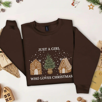 Dark Chocolate brown-colored Beacon & Bramble Christmas sweatshirt featuring the phrase ‘Just a Girl’ above and ‘Who Loves Christmas’ below a festive design of two gingerbread houses and a Christmas tree, surrounded by cozy holiday decor including a wrapped present, a present tag, and greenery.