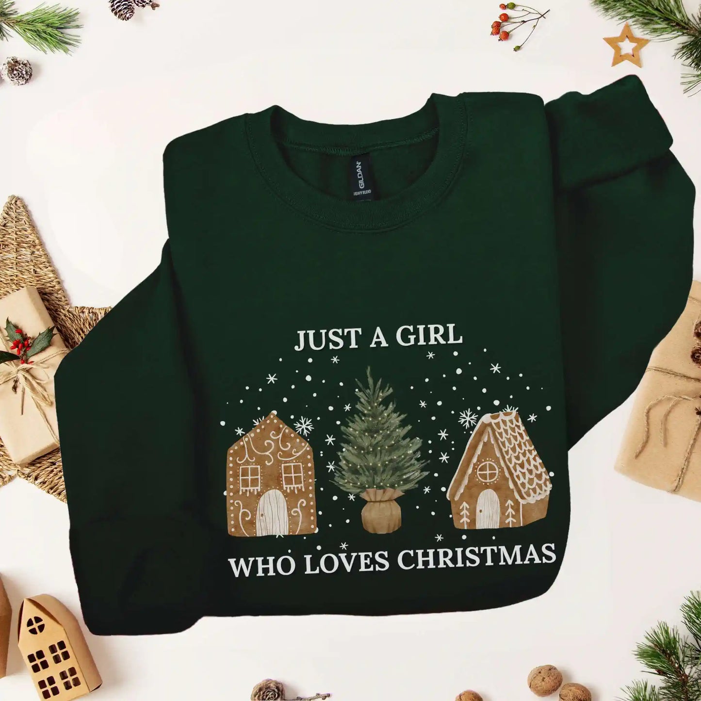 Forest green-colored Beacon & Bramble Christmas sweatshirt featuring the phrase ‘Just a Girl’ above and ‘Who Loves Christmas’ below a festive design of two gingerbread houses and a Christmas tree, surrounded by cozy holiday decor including wrapped presents and greenery.
