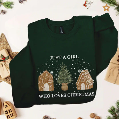 Forest green-colored Beacon & Bramble Christmas sweatshirt featuring the phrase ‘Just a Girl’ above and ‘Who Loves Christmas’ below a festive design of two gingerbread houses and a Christmas tree, surrounded by cozy holiday decor including wrapped presents and greenery.