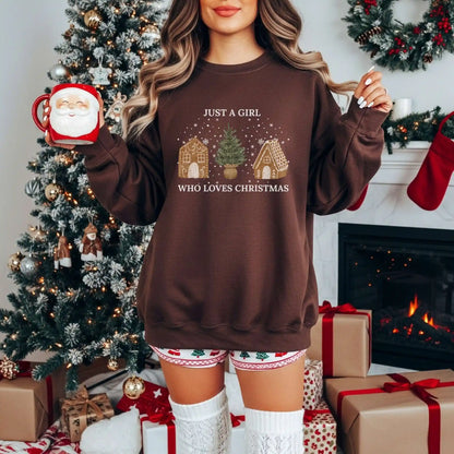 Woman wearing a Dark Chocolate brown Beacon & Bramble Christmas sweatshirt featuring the phrase ‘Just a Girl’ above and ‘Who Loves Christmas’ below a festive design of two gingerbread houses and a Christmas tree. She’s holding a gingerbread-themed mug in a cozy holiday living room with a decorated Christmas tree and wrapped gifts.
