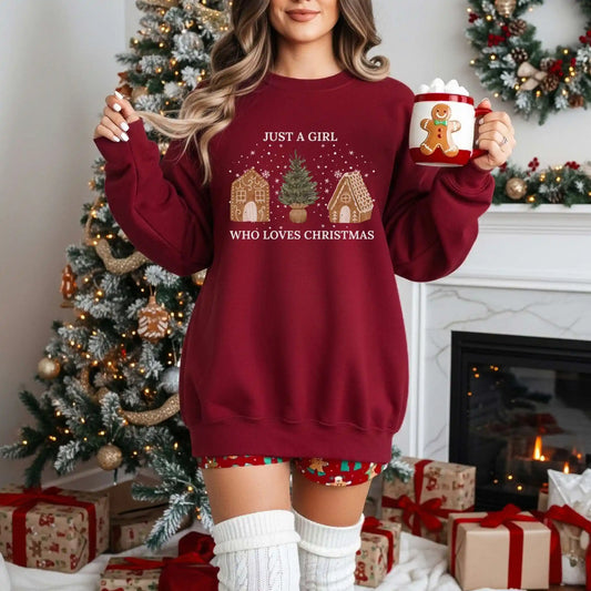 Woman wearing a Garnet red Beacon & Bramble Christmas sweatshirt featuring the phrase ‘Just a Girl’ above and ‘Who Loves Christmas’ below a festive design of two gingerbread houses and a Christmas tree. She’s holding a gingerbread-themed mug in a cozy holiday living room with a decorated Christmas tree and wrapped gifts.