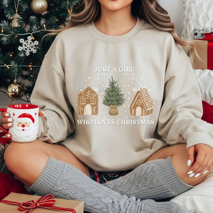 Woman wearing a beige Beacon & Bramble Christmas sweatshirt featuring the phrase ‘Just a Girl’ above and ‘Who Loves Christmas’ below a festive design of two gingerbread houses and a Christmas tree. She’s holding a Santa-themed mug in a cozy holiday living room with a decorated Christmas tree and wrapped gifts.