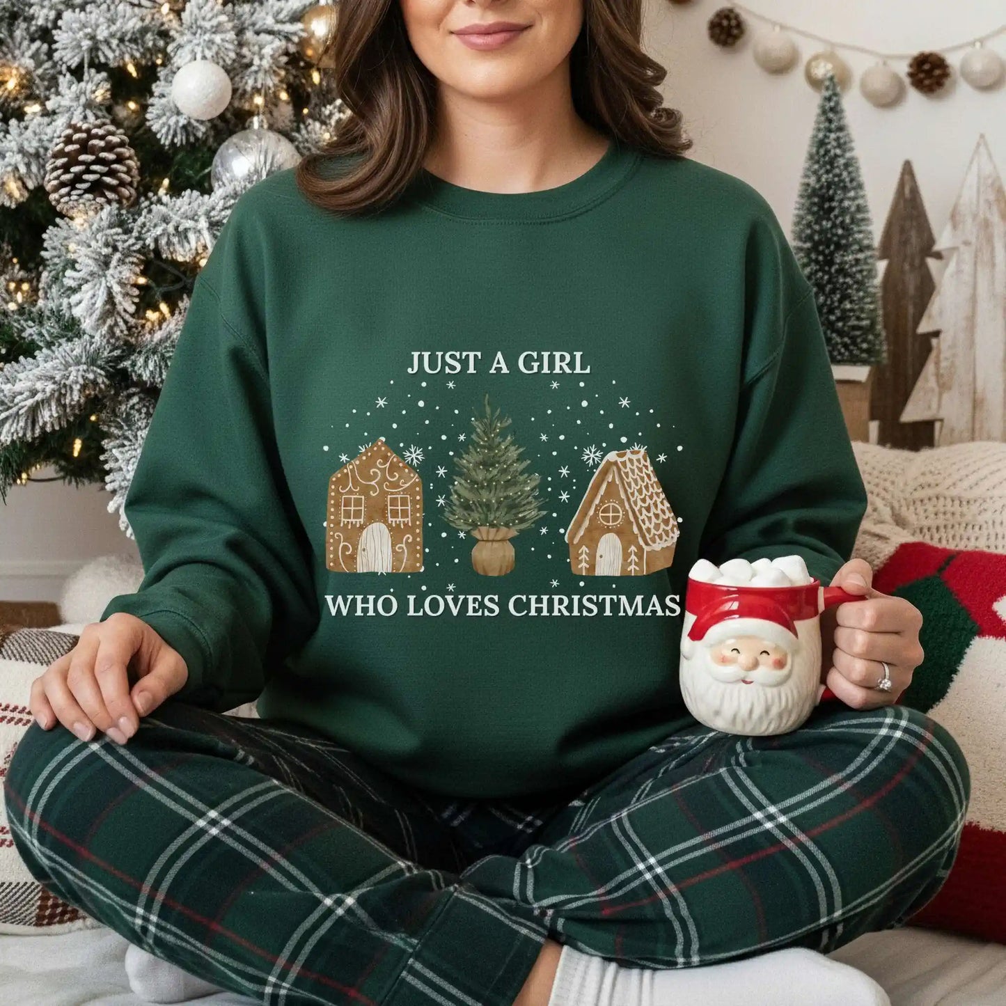 Woman wearing a Forest green Beacon & Bramble Christmas sweatshirt featuring the phrase ‘Just a Girl’ above and ‘Who Loves Christmas’ below a festive design of two gingerbread houses and a Christmas tree. She’s holding a Santa-themed mug in a cozy holiday living room with a decorated Christmas tree.