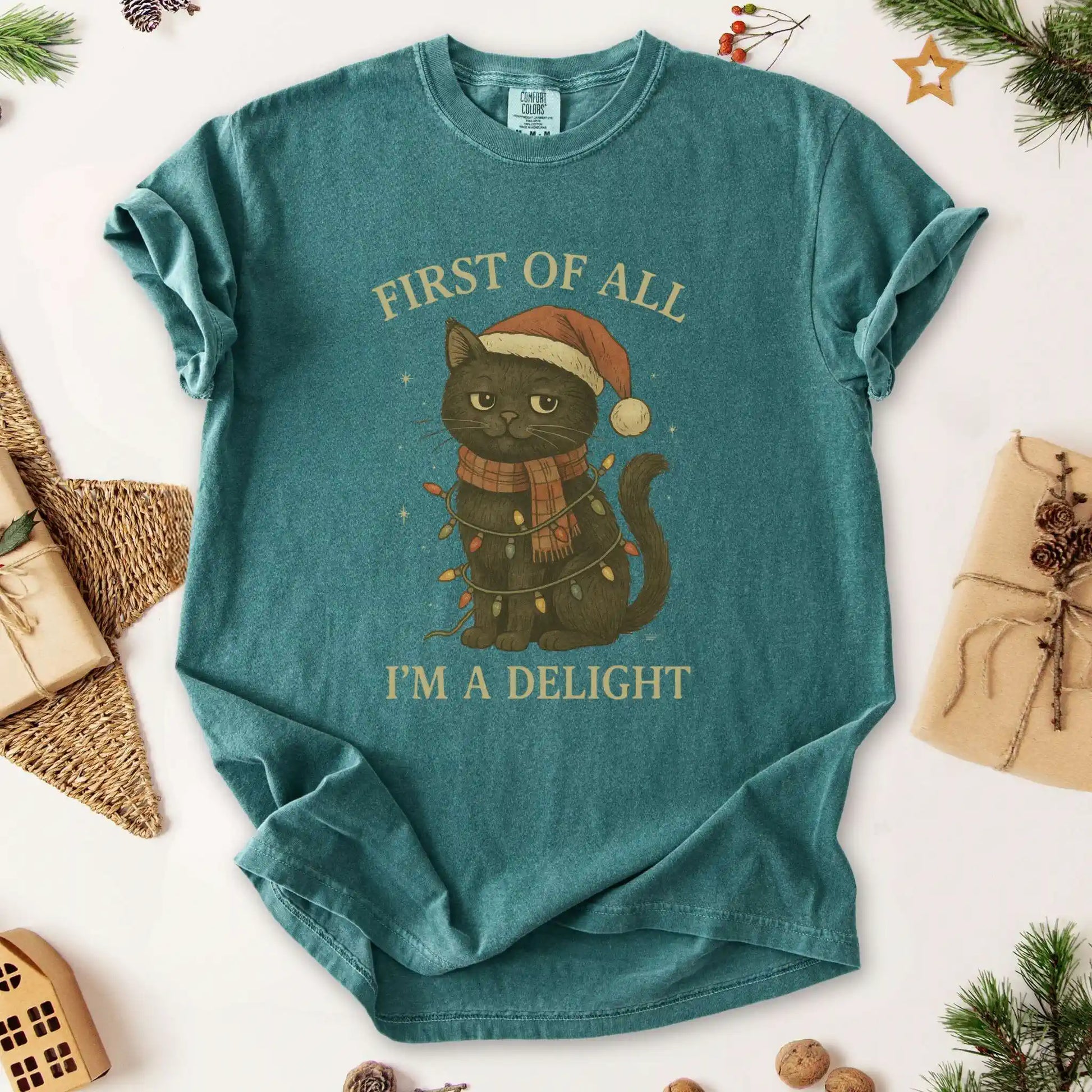Christmas cat graphic t-shirt by Beacon & Bramble featuring a black cat in a Santa hat and scarf wrapped in holiday lights with the phrase ‘First of all, I’m a delight.’ Cozy vintage holiday design on a Blue Spruce green tee.