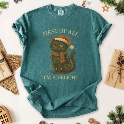 Christmas cat graphic t-shirt by Beacon & Bramble featuring a black cat in a Santa hat and scarf wrapped in holiday lights with the phrase ‘First of all, I’m a delight.’ Cozy vintage holiday design on a Blue Spruce green tee.