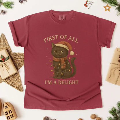 Christmas cat graphic t-shirt by Beacon & Bramble featuring a black cat in a Santa hat and scarf wrapped in holiday lights with the phrase ‘First of all, I’m a delight.’ Cozy vintage holiday design on a red tee.