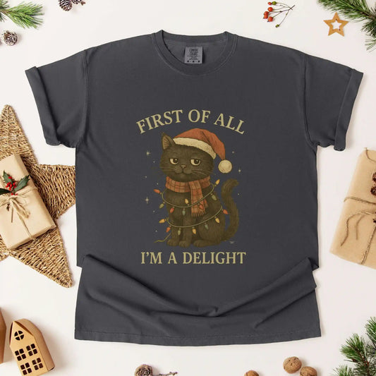 Christmas cat graphic t-shirt by Beacon & Bramble featuring a black cat in a Santa hat and scarf wrapped in holiday lights with the phrase ‘First of all, I’m a delight.’ Cozy vintage holiday design on a dark gray tee.