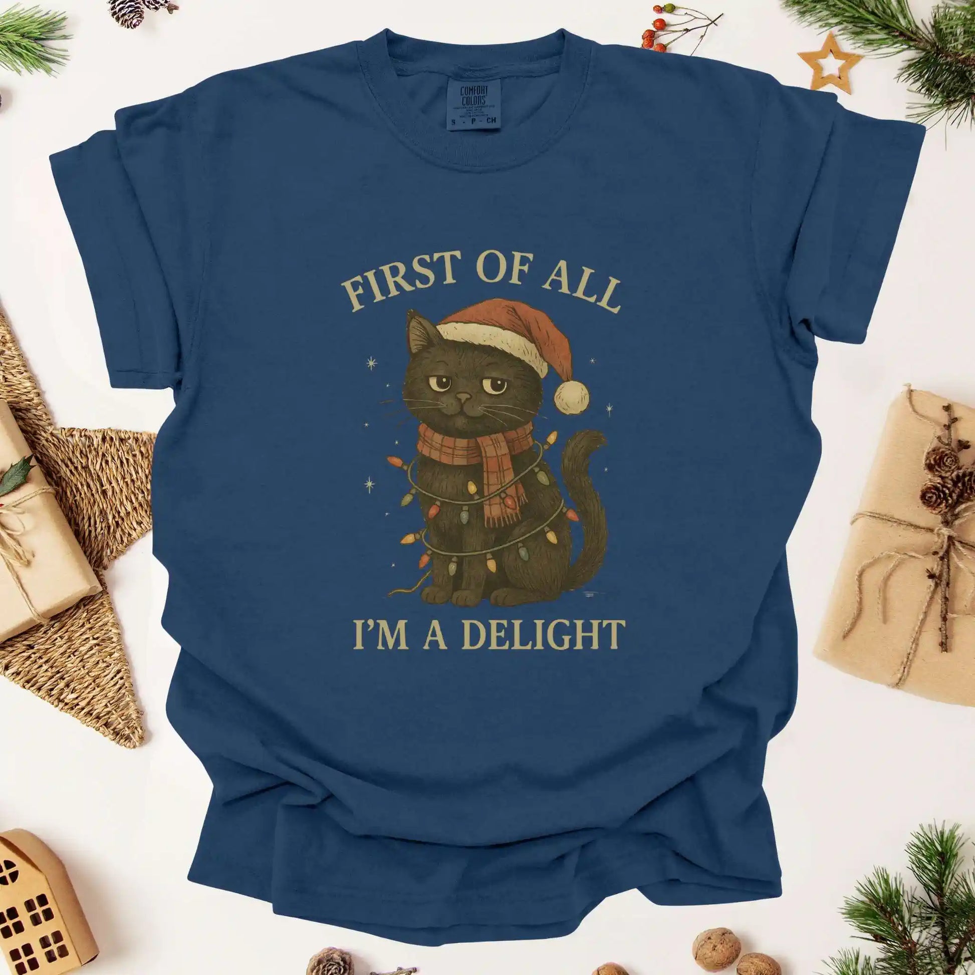 Christmas cat graphic t-shirt by Beacon & Bramble featuring a black cat in a Santa hat and scarf wrapped in holiday lights with the phrase ‘First of all, I’m a delight.’ Cozy vintage holiday design on a navy blue tee.