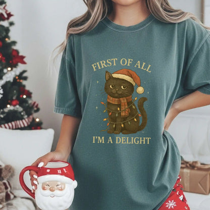 Woman wearing a Blue Spruce green Beacon & Bramble Christmas t-shirt featuring a black cat in a Santa hat and scarf wrapped in holiday lights with the phrase ‘First of all, I’m a delight.’ She’s holding a Santa mug in a cozy holiday living room with a decorated Christmas tree and wrapped gifts.