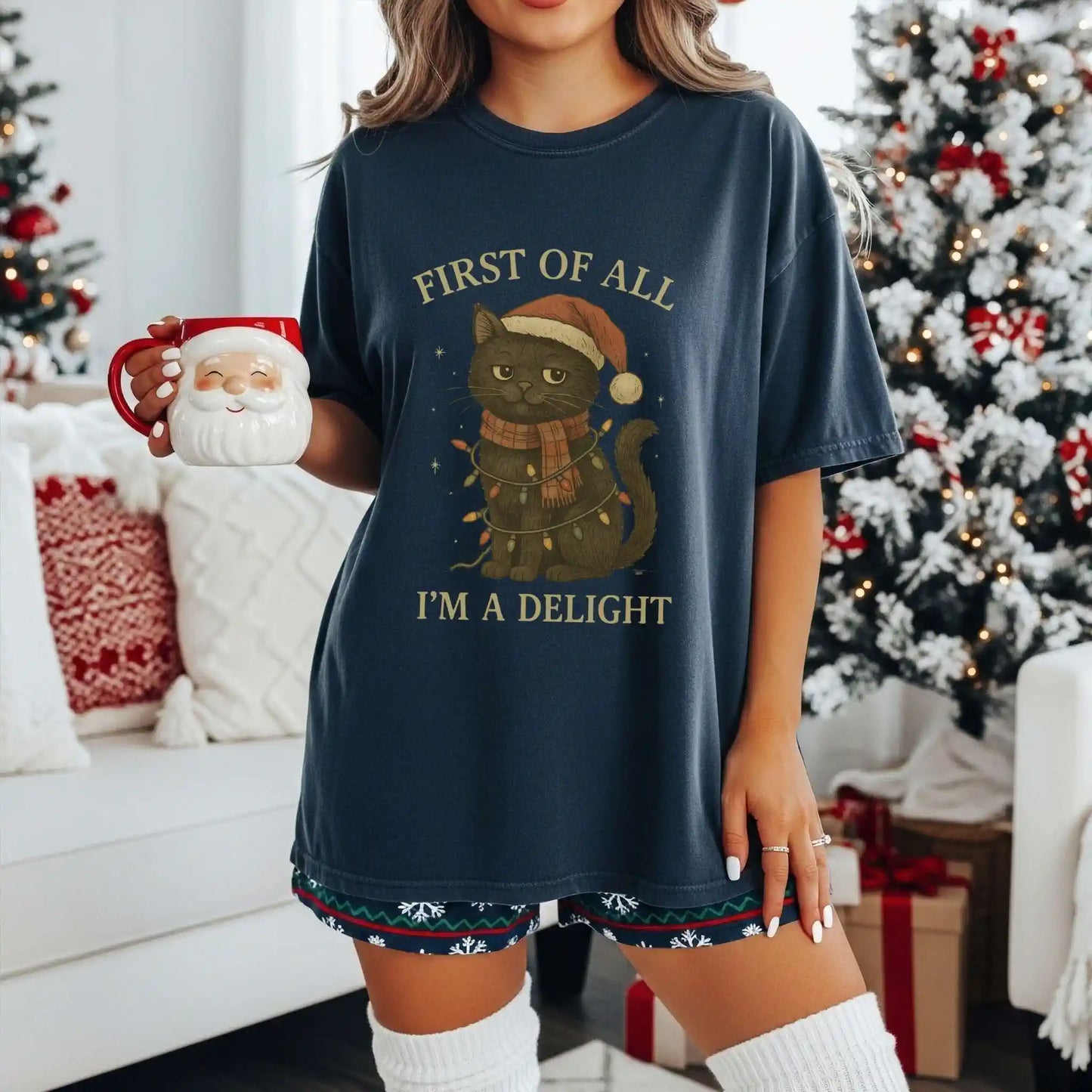 Woman wearing a navy blue Beacon & Bramble Christmas t-shirt featuring a black cat in a Santa hat and scarf wrapped in holiday lights with the phrase ‘First of all, I’m a delight.’ She’s holding a Santa mug in a cozy holiday living room with a decorated Christmas tree and wrapped gifts.