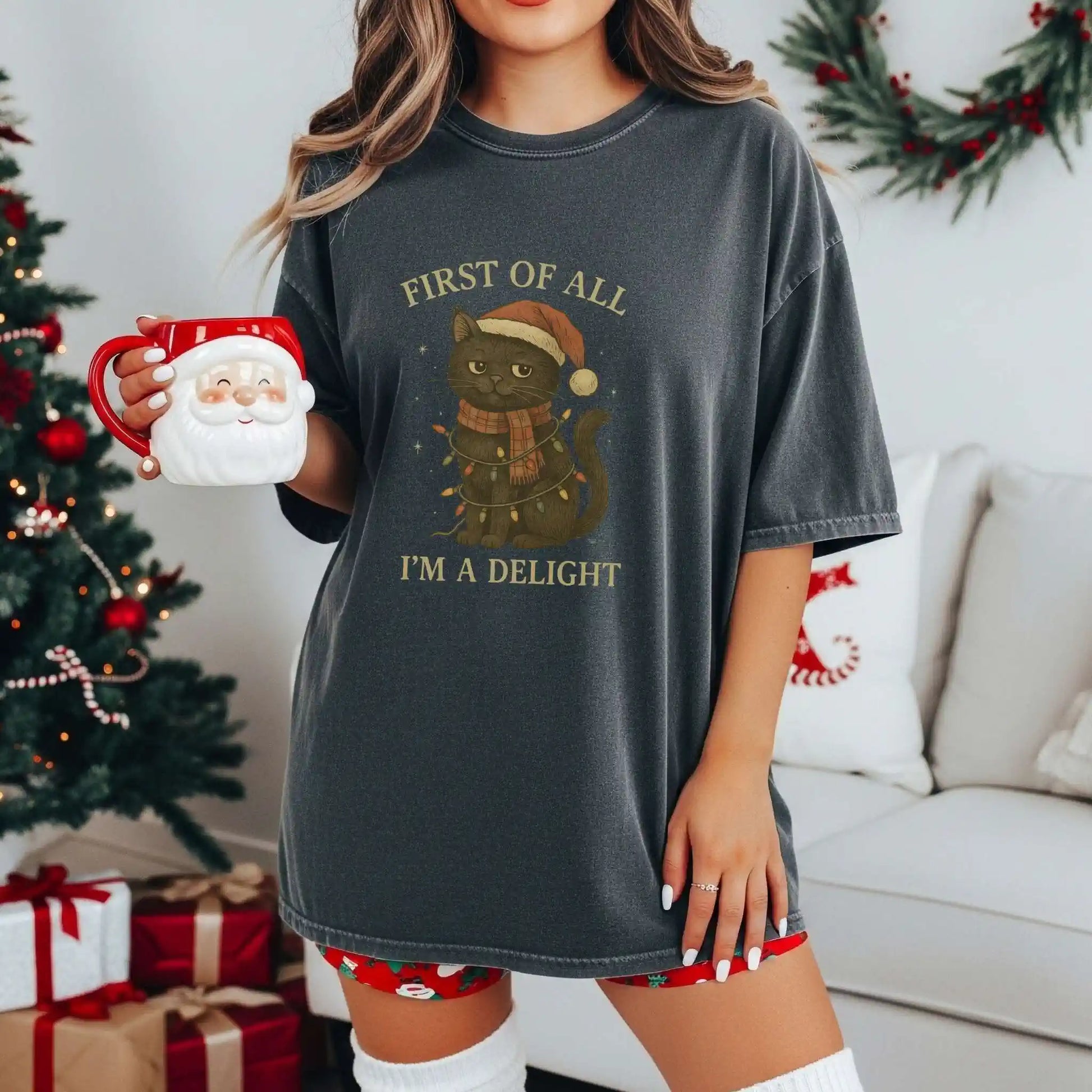 Woman wearing a dark gray Beacon & Bramble Christmas t-shirt featuring a black cat in a Santa hat and scarf wrapped in holiday lights with the phrase ‘First of all, I’m a delight.’ She’s holding a Santa mug in a cozy holiday living room with a decorated Christmas tree and wrapped gifts.