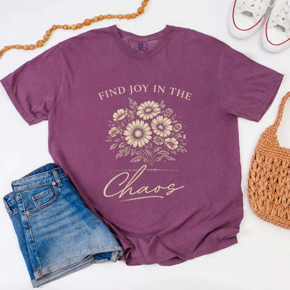 Berry colored t-shirt with floral design and "Find joy in the chaos" text, surrounded by denim shorts, a beaded necklace, and a woven bag.