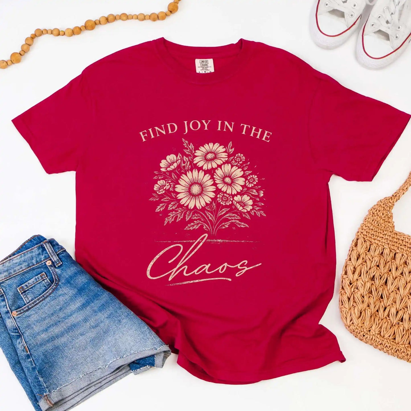 Chili colored t-shirt with floral design and "Find joy in the chaos" text, surrounded by denim shorts, a beaded necklace, and a woven bag.