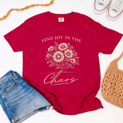 Chili colored t-shirt with floral design and "Find joy in the chaos" text, surrounded by denim shorts, a beaded necklace, and a woven bag.