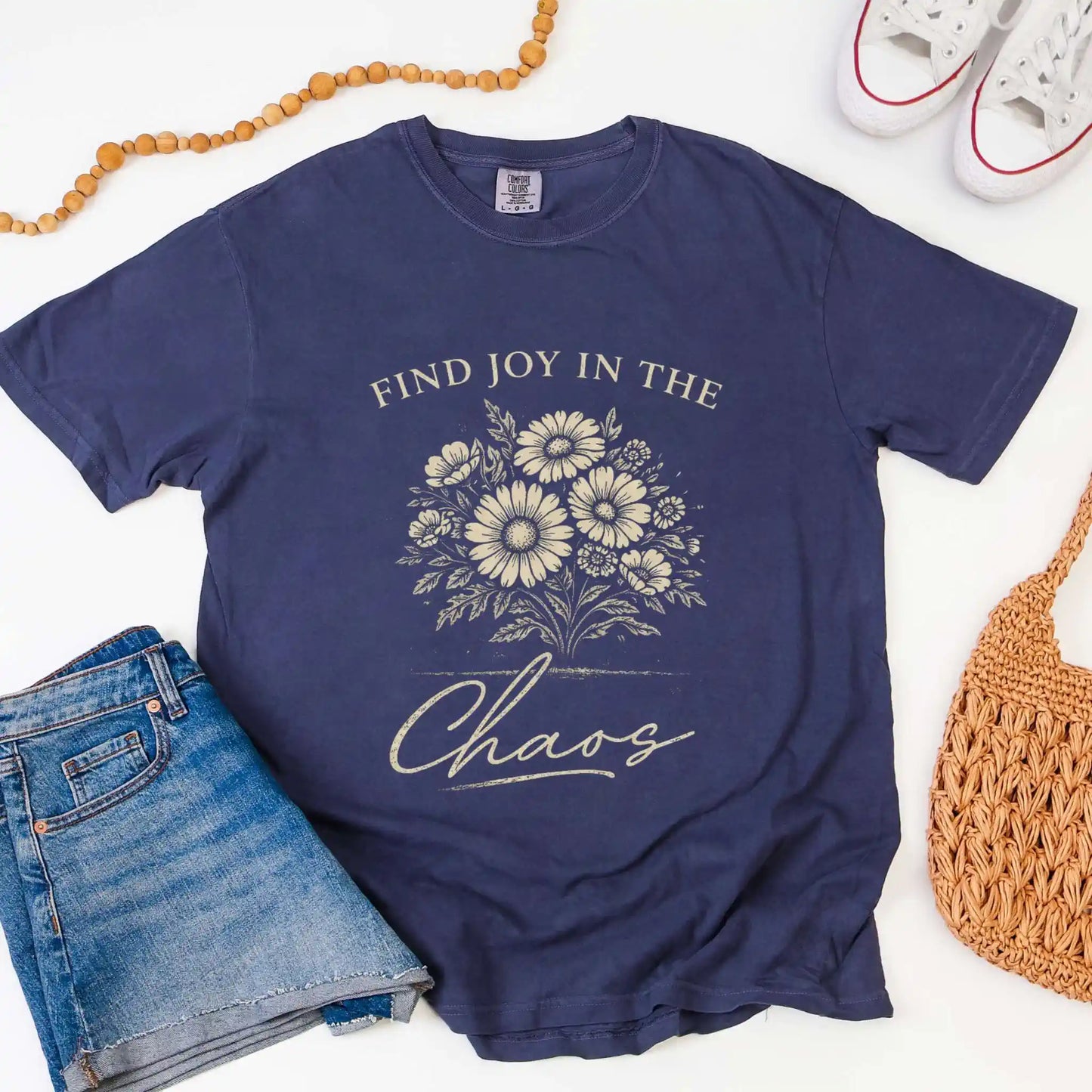 Navy blue t-shirt with floral design and "Find joy in the chaos" text, surrounded by denim shorts, a beaded necklace, and a woven bag.