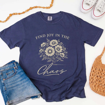Navy blue t-shirt with floral design and "Find joy in the chaos" text, surrounded by denim shorts, a beaded necklace, and a woven bag.