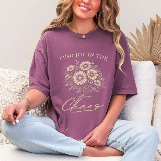 Woman wearing a Berry colored t-shirt with floral design and "Find joy in the chaos" text, sitting on a couch.