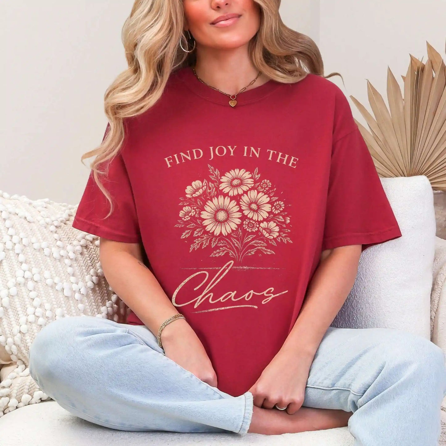 Woman wearing a Chili colored t-shirt with floral design and "Find joy in the chaos" text, sitting on a couch.