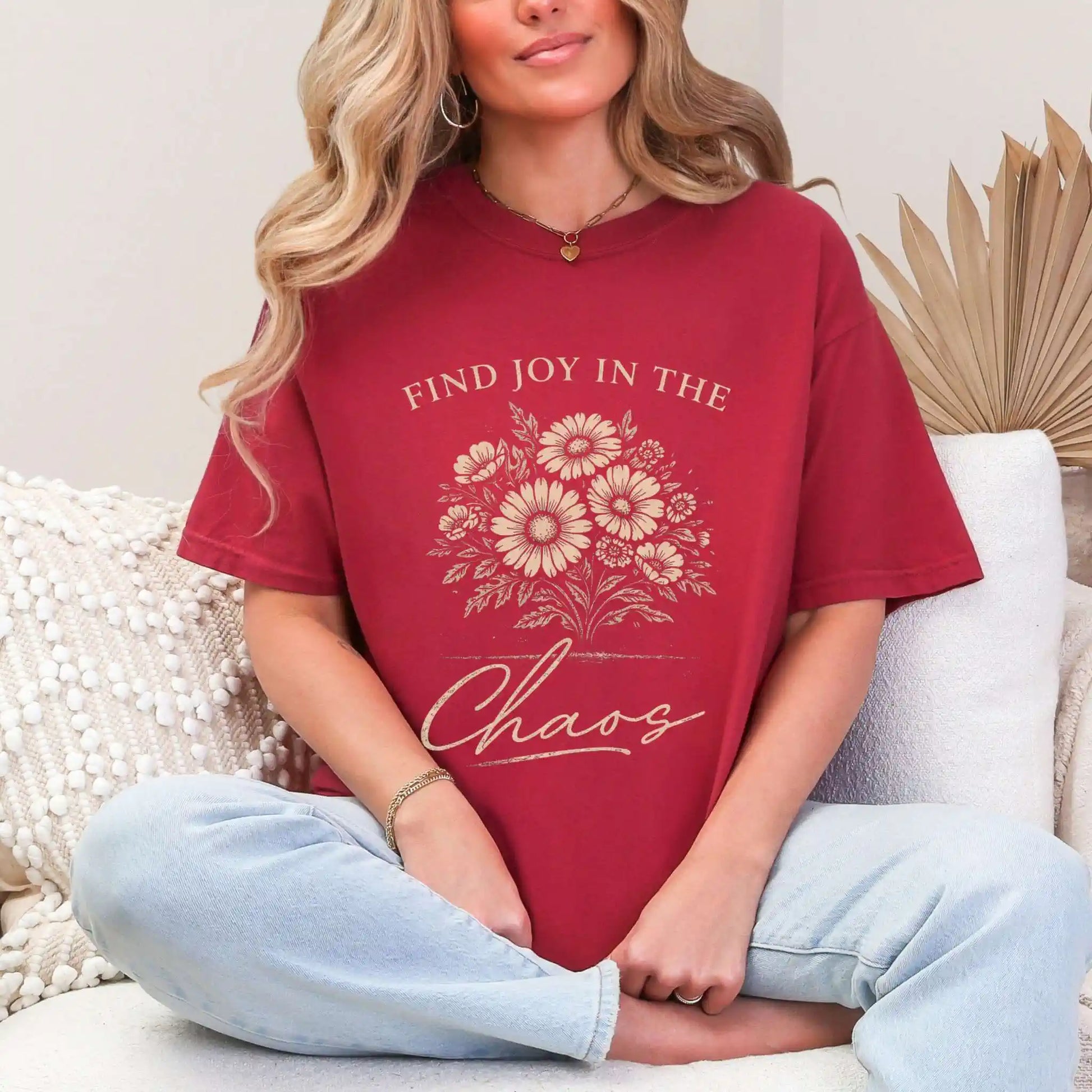 Woman wearing a Chili colored t-shirt with floral design and "Find joy in the chaos" text, sitting on a couch.