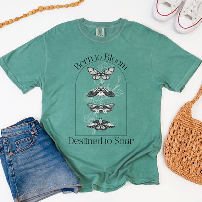 Green t-shirt with butterfly design and 'Born to Bloom, Destined to Soar' text, paired with denim shorts and a woven bag on a white background.